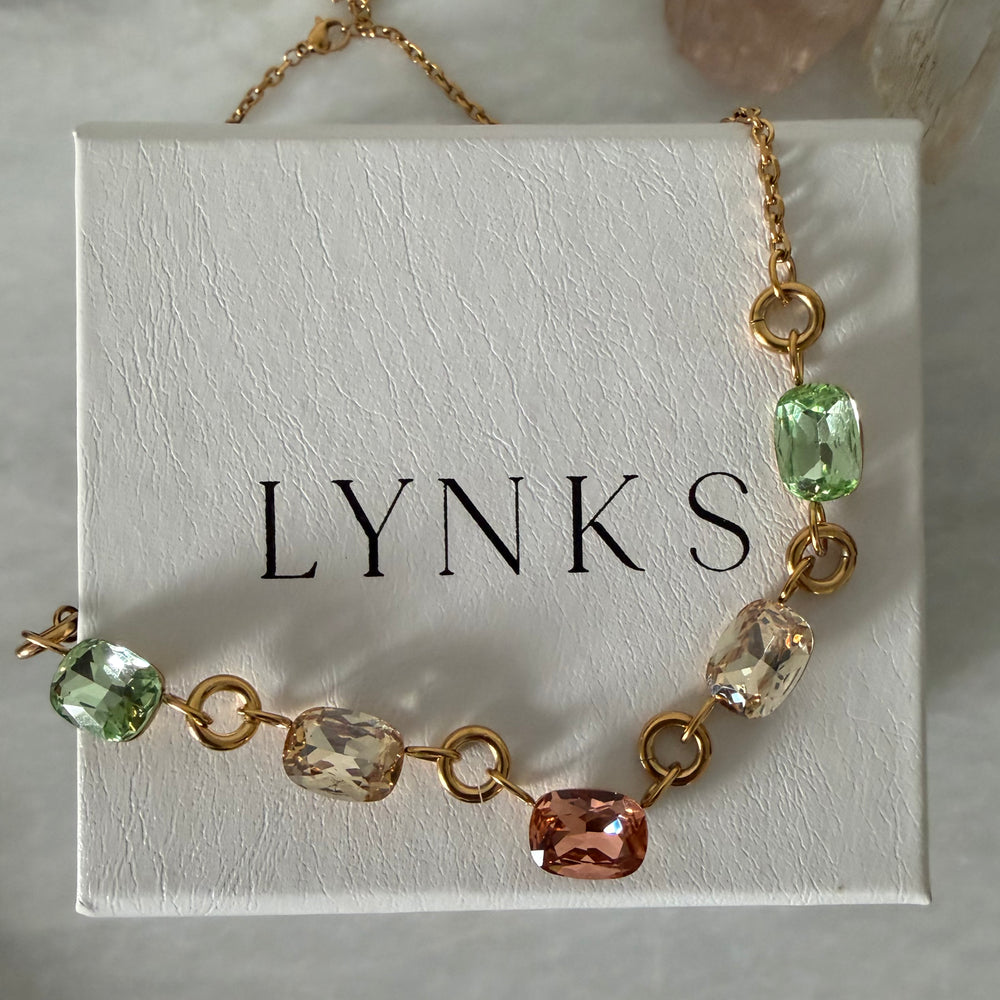 A gold chain bracelet with faceted gemstone links in green, peach, and clear, displayed on a white textured jewelry box printed with the brand name 'LYNKS'.