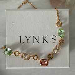 A gold chain bracelet with faceted gemstone links in green, peach, and clear, displayed on a white textured jewelry box printed with the brand name 'LYNKS'.