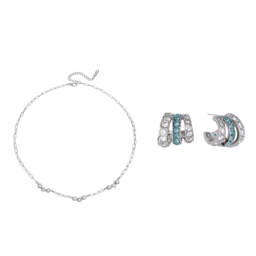 A jewelry set featuring a delicate silver chain bracelet and matching silver huggie hoop earrings, both adorned with small light blue stones, displayed on a clean white background.